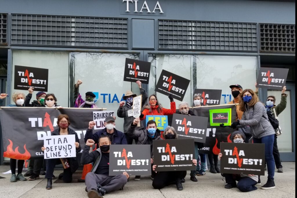 A group of about a dozen people fists raised, wearing masks in front of TIAA's building with signs that read "TIAA Divest" and "Defund Line 3"