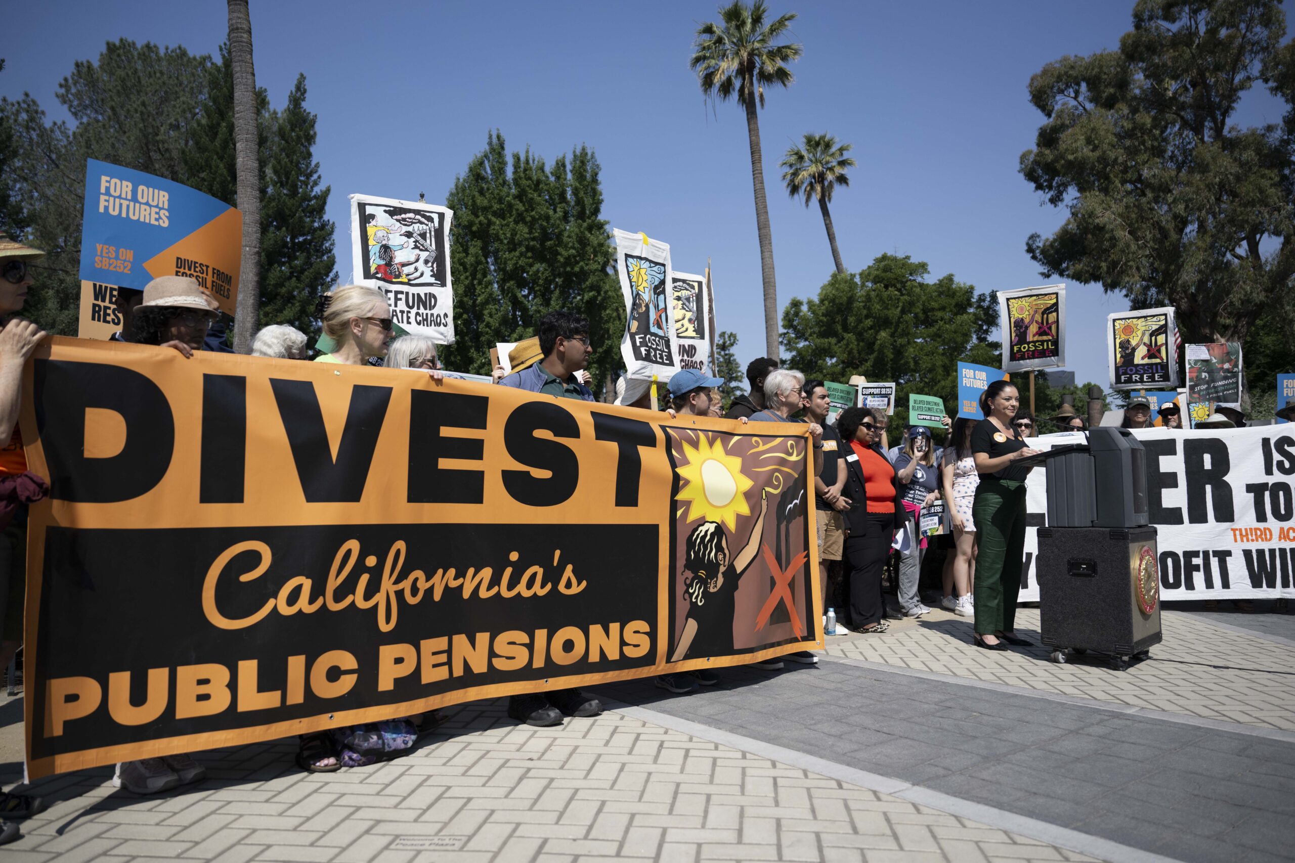 Group of Californians at a press conference behind an orange banner reading "Divest California's Public Pensions"